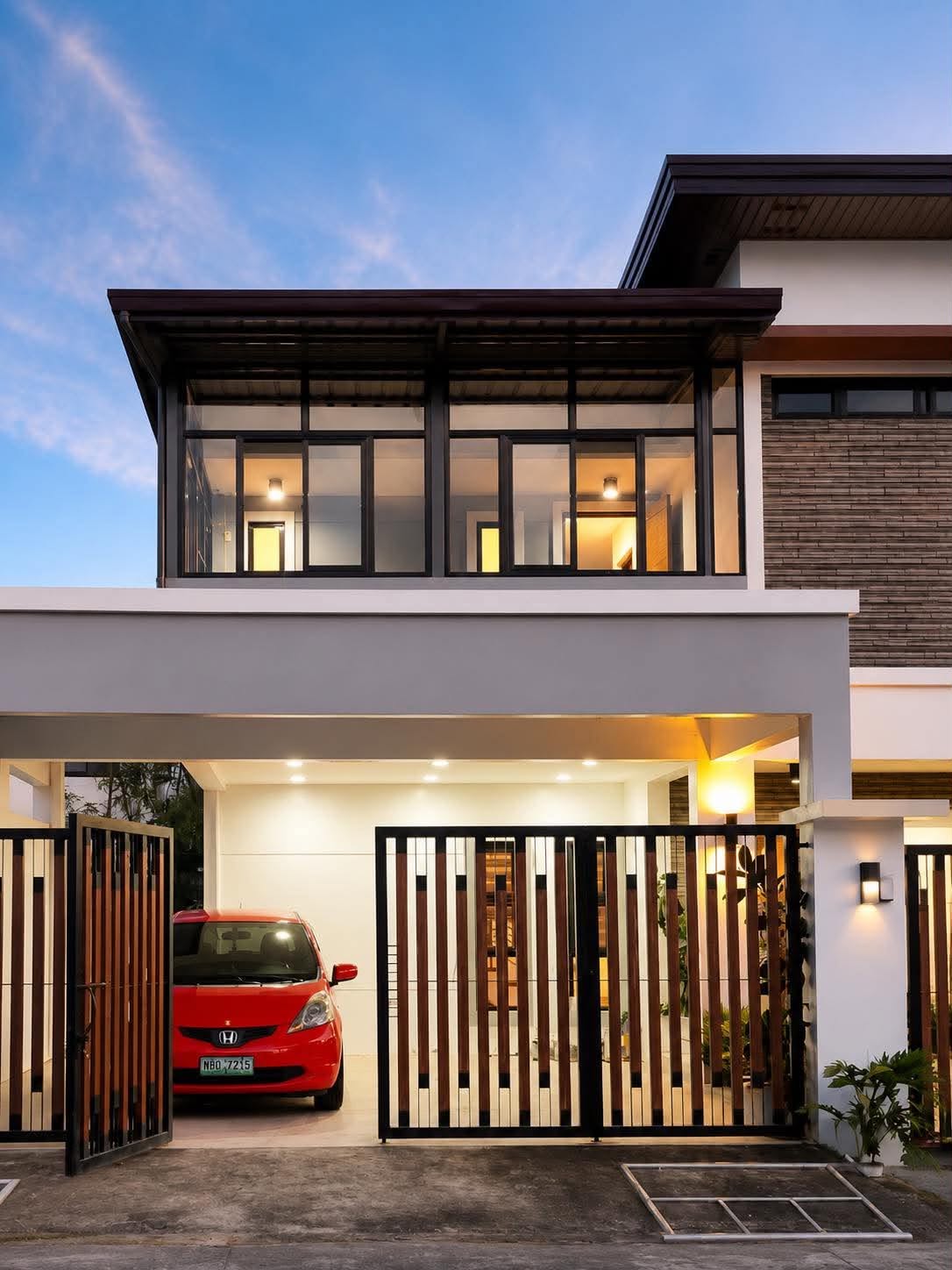 A modern two-storey house at dusk, with a timber slat gate, stone cladding and a lit carport