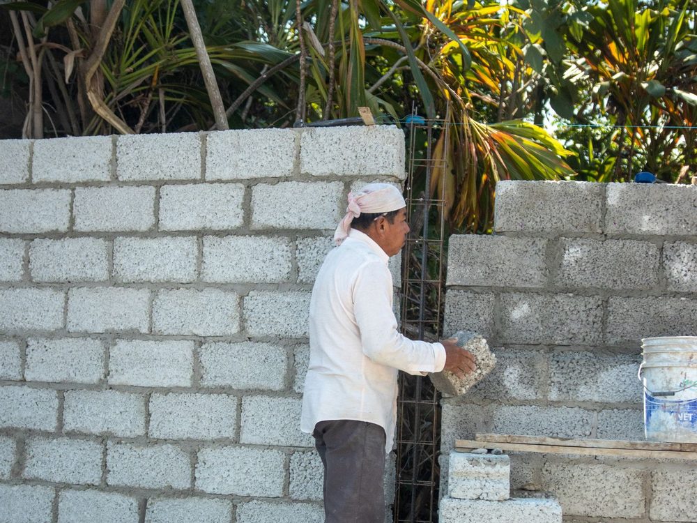 A mason laying concrete hollow block against a reinforced column, tropical planting behind the wall